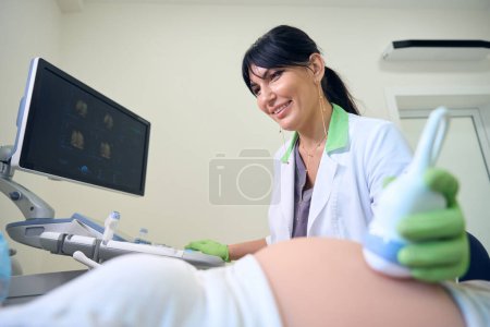 Photo for Smiling adult european female gynecologist doing ultrasound examination of cropped pregnant woman in clinic. Concept of pregnancy examination - Royalty Free Image