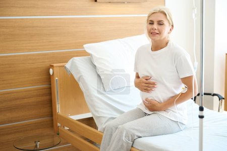 Photo for Thoughtful adult european pregnant woman sitting on medical bed and embracing her belly in maternity hospital - Royalty Free Image