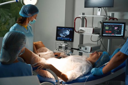 Foto de Personal del hospital que mira el monitor durante el examen por ultrasonido de la mujer embarazada caucásica en equipos modernos antes del parto en la clínica reproductiva. Embarazo y maternidad - Imagen libre de derechos