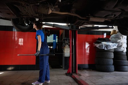 Photo for Female in blue uniform stands under the car, raised on a lift, the foreman has a crowbar in his hands - Royalty Free Image
