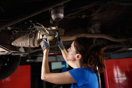 Téléchargez les photos : Mécanicien automobile féminin inspecte le châssis de la voiture, soulevé sur un ascenseur, le maître a des outils dans ses mains - en image libre de droit