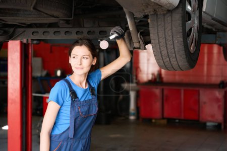 Foto de Especialista en reparación de automóviles se encuentra debajo de un coche levantado en un ascensor, una mujer en un uniforme azul - Imagen libre de derechos