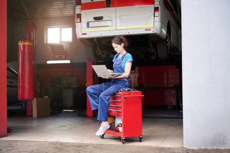 Téléchargez les photos : Spécialiste de la réparation automobile est assis avec un ordinateur portable près d'une voiture soulevée sur un ascenseur, une femme dans un uniforme bleu - en image libre de droit