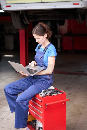 Photo for Woman sits with laptop on red stand near a car raised on a lift, a woman in a blue uniform - Royalty Free Image