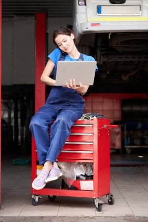 Téléchargez les photos : Femme mécanicien automobile assis avec un ordinateur portable sur un stand rouge, elle communique sur un téléphone mobile - en image libre de droit