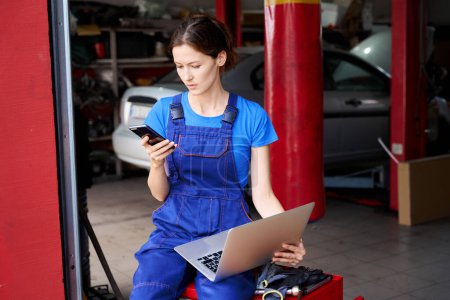 Téléchargez les photos : Mécanicien automobile féminine avec un ordinateur portable dans un atelier de réparation automobile, elle communique sur un téléphone portable - en image libre de droit