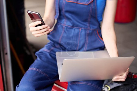 Téléchargez les photos : Mécanicien automobile assis avec un ordinateur portable et téléphone portable sur une armoire rouge, une femme travaille dans un atelier de réparation automobile - en image libre de droit