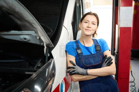 Photo for Nice female auto mechanic in overalls stands near a specialized car, she is wearing protective gloves - Royalty Free Image