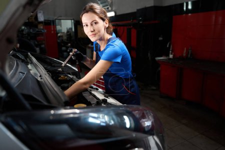 Téléchargez les photos : Jeune femme travaille sous le capot ouvert d'une voiture, elle a un outil spécial dans ses mains - en image libre de droit