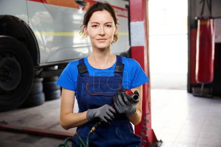 Photo for Female is standing in an auto repair shop with a pneumatic wrench, she is wearing blue overalls - Royalty Free Image