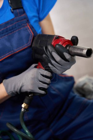 Photo for Auto mechanic in a car repair shop with a pneumatic wrench, she is in a blue work overall - Royalty Free Image