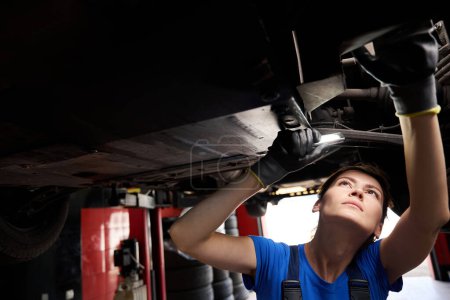 Téléchargez les photos : Jeune technicien de réparation inspectant le dessous d'une voiture dans un atelier de réparation automobile, femme à l'aide d'une lampe de poche - en image libre de droit
