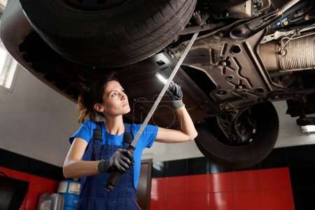 Photo for Woman in a blue uniform inspects a car raised on a lift, the woman uses a special tool - Royalty Free Image