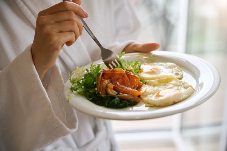 Téléchargez les photos : Photo recadrée de femme en peignoir piquant salade avec fourchette sur l'assiette dans sa main - en image libre de droit