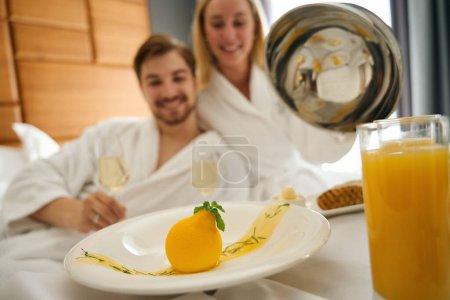 Photo for Woman and man enjoying champagne breakfast, table set on large bed - Royalty Free Image