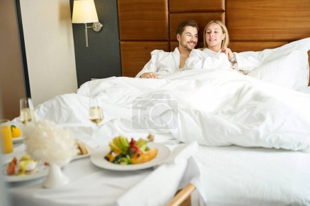 Photo for Blonde woman and her boyfriend are relaxing in a hotel on a large bed, breakfast with champagne is served nearby - Royalty Free Image