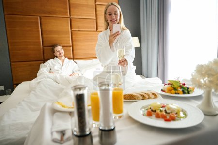 Foto de Pareja en la sala de recién casados se sirve el desayuno con champán en la cama, una mujer toma fotografías de la mesa servida - Imagen libre de derechos