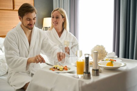 Photo for Young couple communicates over breakfast in the honeymoon room, the woman has a glass of champagne in her hands - Royalty Free Image