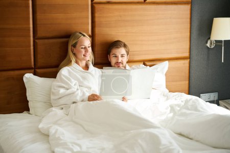 Photo for Waking up guy and girlfriend study the menu in bed, the couple chooses breakfast in the room - Royalty Free Image