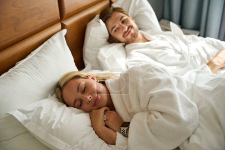 Téléchargez les photos : Jeune homme se trouve à côté d'une blonde endormie, un couple repose sur un grand lit - en image libre de droit