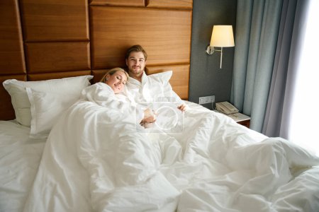 Téléchargez les photos : Beau couple se trouve étreignant sur des oreillers doux dans une chambre d'hôtel, les gens en peignoirs moelleux - en image libre de droit