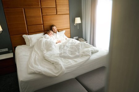 Photo for Woman and a man lie hugging on a large bed, they are wearing hotel terry robes - Royalty Free Image