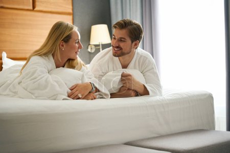 Téléchargez les photos : Femme et un homme communiquent tout en étant couchés sur un grand lit, ils portent des robes éponge hôtel - en image libre de droit