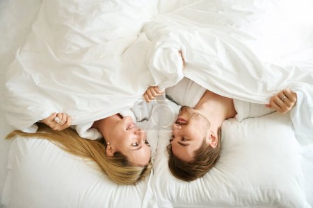 Photo for Young couple lying in bed in a hotel room, they look at each other lovingly - Royalty Free Image