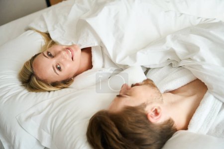Téléchargez les photos : Heureux homme et femme couchés dans leur lit, ils sont dans des robes douillettes éponge - en image libre de droit