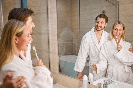 Téléchargez les photos : Épouses gaies dans la salle de bain faisant leur routine du matin, un couple dans des robes éponge confortables - en image libre de droit
