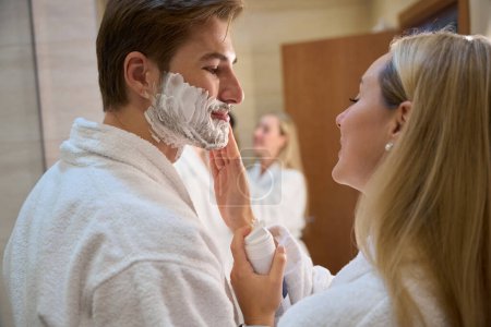 Foto de Mujer rubia aplica espuma de afeitar a los maridos mejillas, pareja haciendo rutina de la mañana en el baño - Imagen libre de derechos