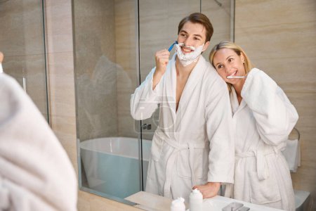 Photo for Happy couple in bathroom doing morning routine, man shaving, woman brushing teeth - Royalty Free Image