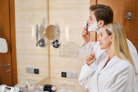 Foto de Esposo y esposa en el baño haciendo su rutina de la mañana, hombre afeitándose, mujer cepillándose los dientes - Imagen libre de derechos