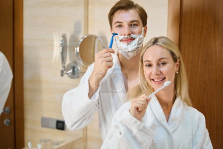 Foto de Pareja joven en el baño haciendo su rutina matutina, el hombre afeitándose, la mujer cepillándose los dientes - Imagen libre de derechos