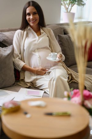 Foto de Encantadora mujer embarazada se sienta en el sofá con una taza de café, bocetos de ropa se encuentran junto a ella - Imagen libre de derechos