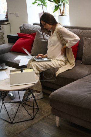 Téléchargez les photos : Designer avec une mauvaise santé travaille sur un ordinateur portable, elle est assise sur un grand canapé - en image libre de droit