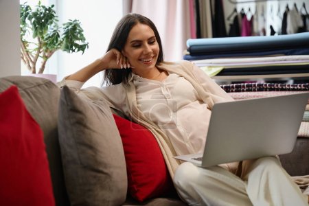Téléchargez les photos : Femme enceinte s'assoit confortablement dans un espace de détente avec un ordinateur portable, elle a un ventre rond mignon - en image libre de droit