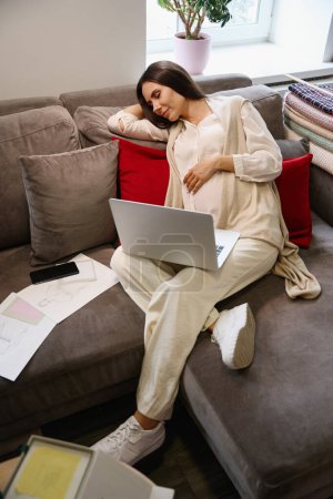Téléchargez les photos : Femme fatiguée est somnoler sur des oreillers doux avec ordinateur portable dans ses mains, à côté d'elle sont des croquis de travail de vêtements - en image libre de droit