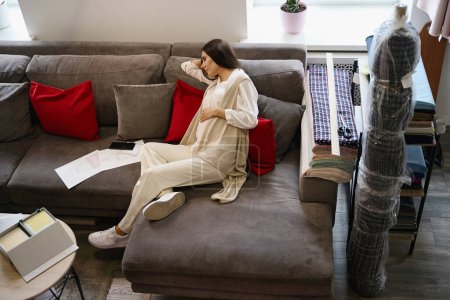 Téléchargez les photos : Femme enceinte est somnoler sur des oreillers doux dans un espace de détente, à côté d'elle sont des croquis de travail de vêtements - en image libre de droit