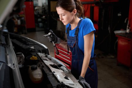 Téléchargez les photos : Femme en bleu vêtements de travail inspecte le moteur sous le capot ouvert, elle a un outil spécial dans ses mains - en image libre de droit