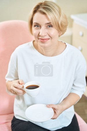 Foto de Retrato de mujer caucásica adulta sonriente con taza de té o café mirando a la cámara en el salón de belleza borrosa - Imagen libre de derechos