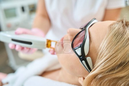 Photo for Partial image of cosmetologist doing laser epilation with IPL photorejuvenation device of woman face wearing glasses in blurred beauty salon. Concept of face skin care - Royalty Free Image