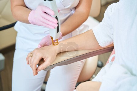 Photo for Partial image of female beautician doing laser epilation with IPL photorejuvenation device of woman hand in blurred beauty salon. Concept of body skin care - Royalty Free Image