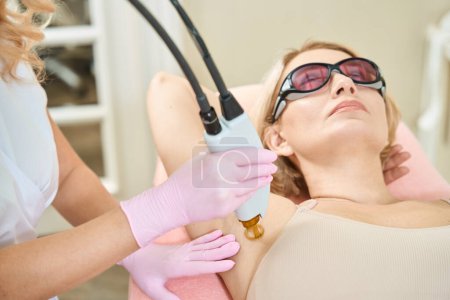 Photo for Partial image of female beautician doing laser epilation with IPL photorejuvenation device of armpit of adult caucasian woman wearing glasses in blurred beauty salon. Concept of body skin care - Royalty Free Image
