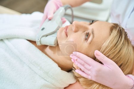Photo for Partial female beautician doing laser depilation with laser hair removal machine of face of adult caucasian woman in blurred beauty salon. Concept of face skin care - Royalty Free Image