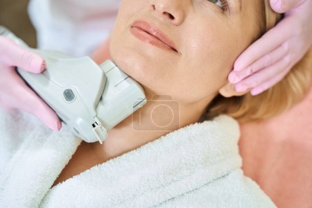 Photo for Obscure face if adult woman during partial female beautician doing laser depilation with laser hair removal machine to her in blurred beauty salon. Concept of face skin care - Royalty Free Image