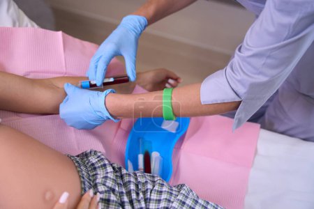 Téléchargez les photos : Image recadrée d'une infirmière prenant du sang avec une seringue de femme enceinte sur un lit médical à l'hôpital. Concept de grossesse et de maternité - en image libre de droit