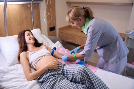 Téléchargez les photos : Infirmière européenne faisant une injection avec une seringue de jeune femme enceinte caucasienne souriante sur un lit médical à l'hôpital. Concept de grossesse et de maternité - en image libre de droit