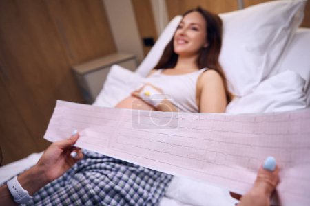 Téléchargez les photos : Concentration sélective de l'électrocardiogramme dans les mains d'une infirmière partielle près d'une patiente enceinte floue sur un lit médical à l'hôpital. Concept de grossesse et de maternité - en image libre de droit