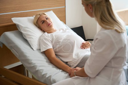 Téléchargez les photos : Infirmière tenant la main d'une femme enceinte caucasienne adulte sur un lit médical à la maternité - en image libre de droit